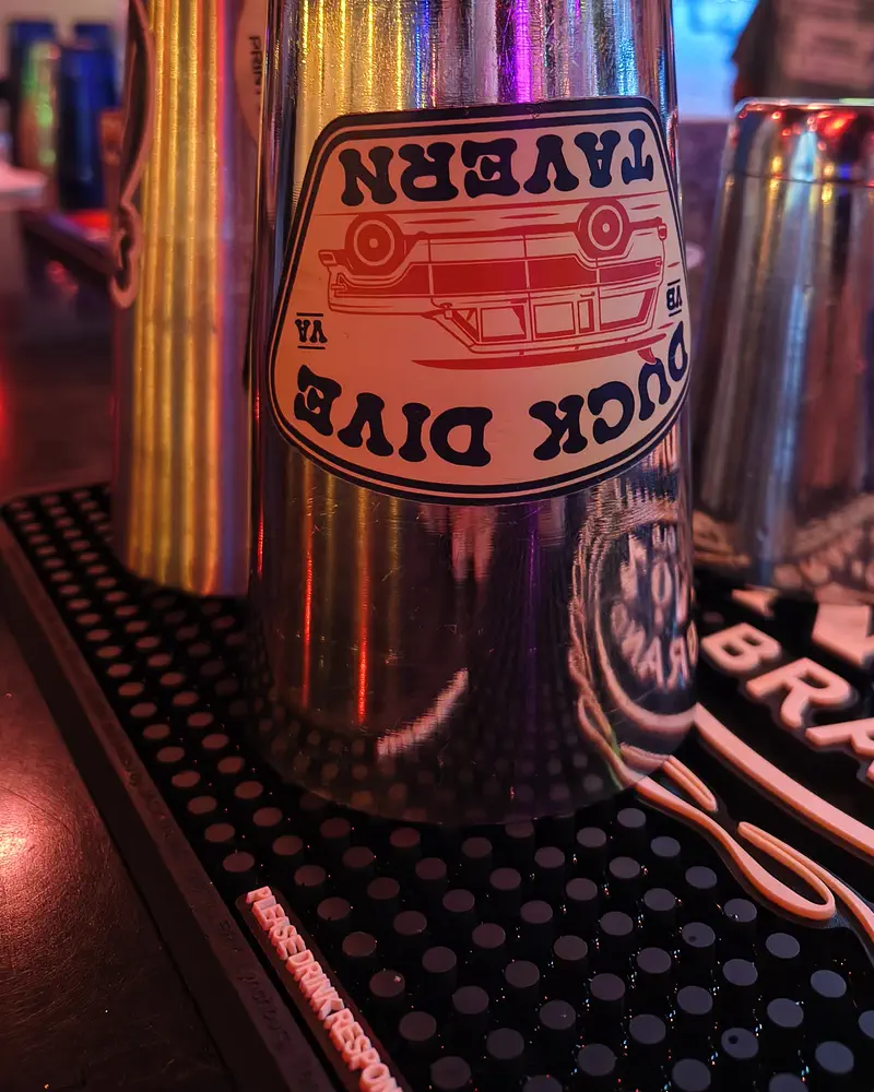 Steel shaker tins on the bar mat, one with the Duck Dive badge sticker, the neon-lit bar behind