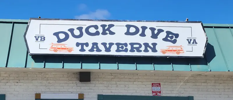 The Duck Dive Tavern street sign with the VB and VA boxes and two woody wagons, on a teal metal roof under a blue sky